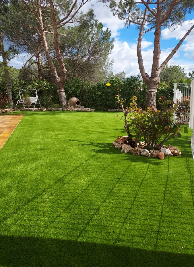 Jardín con césped artificial, pinos y una zona de plantas rodeada de piedras